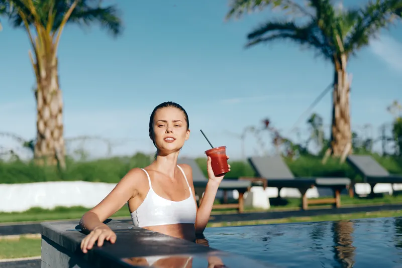 A young woman in a white top leans on the pool edge, holding a red smoothie with a straw, enjoying a bright casual lifestyle moment by the water on a sunny day.