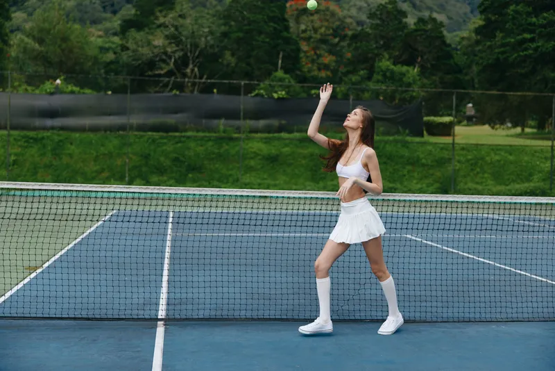 Young female tennis player serving on a court, wearing a sporty white outfit and showing determination against a backdrop of green trees and blue sky