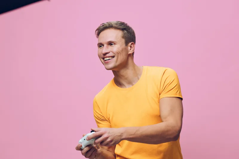 Smiling man playing video game, holding game controller, casual yellow t-shirt, cheerful expression, indoor studio setting, pink background, entertainment lifestyle concept