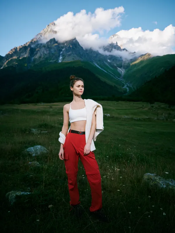 Mountain landscape with a stylish woman standing in a green meadow, wearing a white crop top and red pants, jacket draped over shoulder, crisp sky and distant peaks.