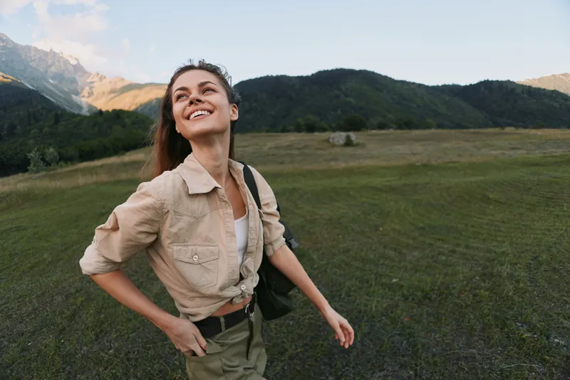 Young woman explores a grassy plain near mountains, smiling softly as warm light fills the landscape and she moves freely, enjoying nature and fresh air.