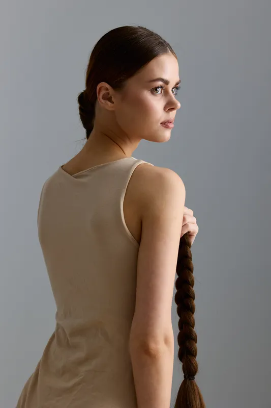 Young woman with long braided hair wearing a beige sleeveless top stands thoughtfully against a neutral gray background in soft natural light.