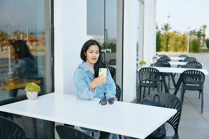 Woman at a cafe outdoor terrace using smartphone, relaxed and focused, enjoying a sunny moment in a modern casual setting with bright seating and glass windows.