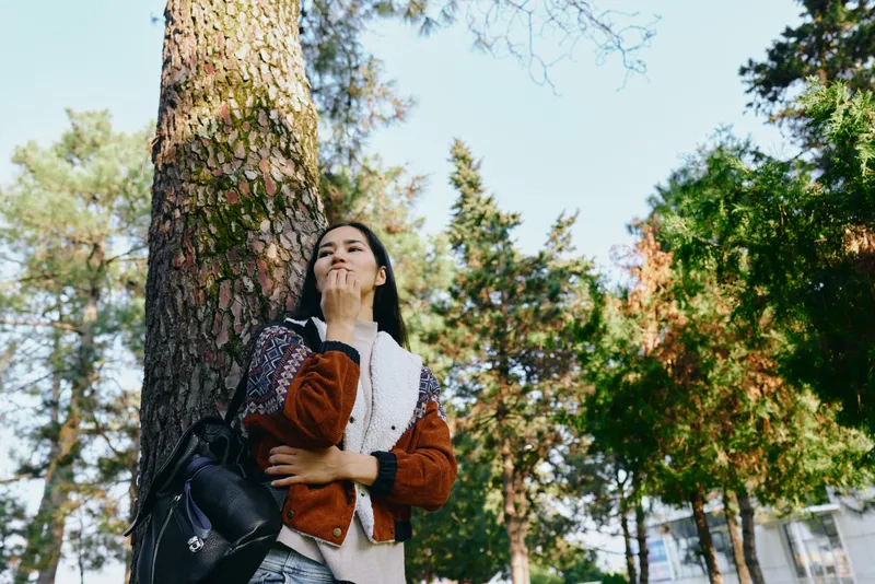 Woman stands near a tall tree in a park, enjoying sunlight, greenery and a light breeze, wearing a jacket and backpack, a casual pose that captures outdoor leisure and nature