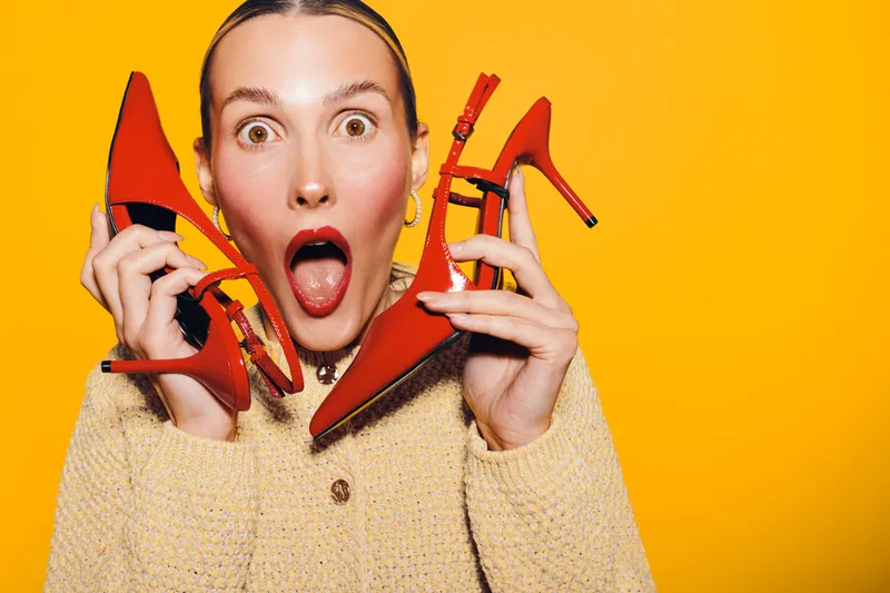 Surprised woman holds red high heels near face on yellow background. Concept of fashion, style, footwear, and shopping excitement with bright studio lighting and casual outfit.