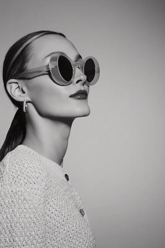 Elegant caucasian woman with fair skin wearing round sunglasses and knitted top poses in vintage style with confident expression.