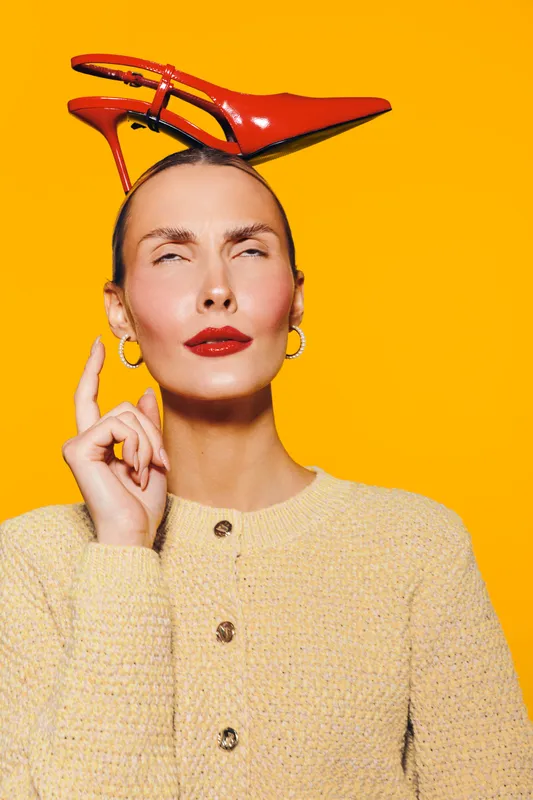 Woman with red high heel shoe on head poses with thoughtful expression against bright yellow background. Fashion concept with bold makeup and creative styling.