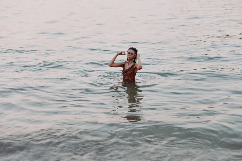 Woman standing alone in calm water wearing a red dress, expressing contemplation and relaxation in a natural outdoor setting.