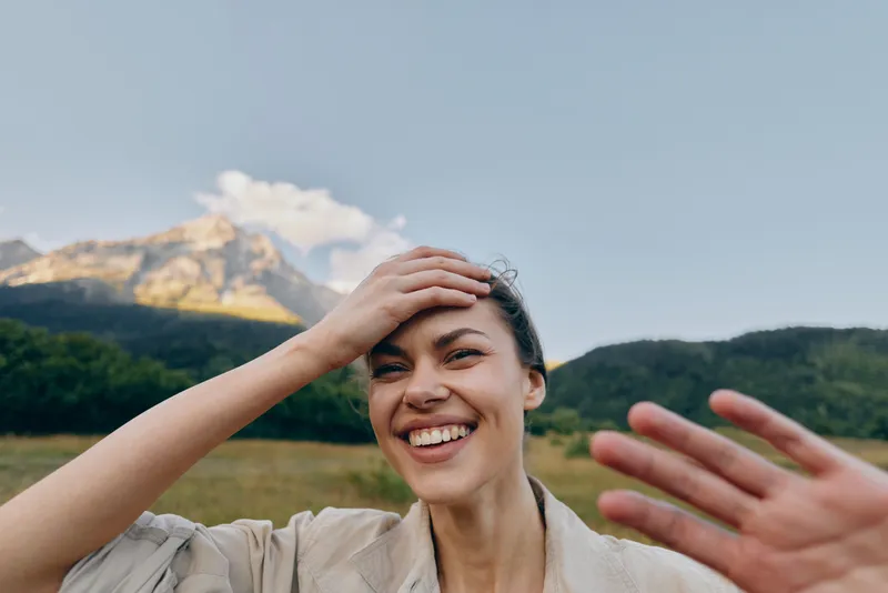 A happy woman outdoors smiles broadly while taking a selfie with mountains and forest in the background. Concept for lifestyle, nature, and joy in everyday moments.