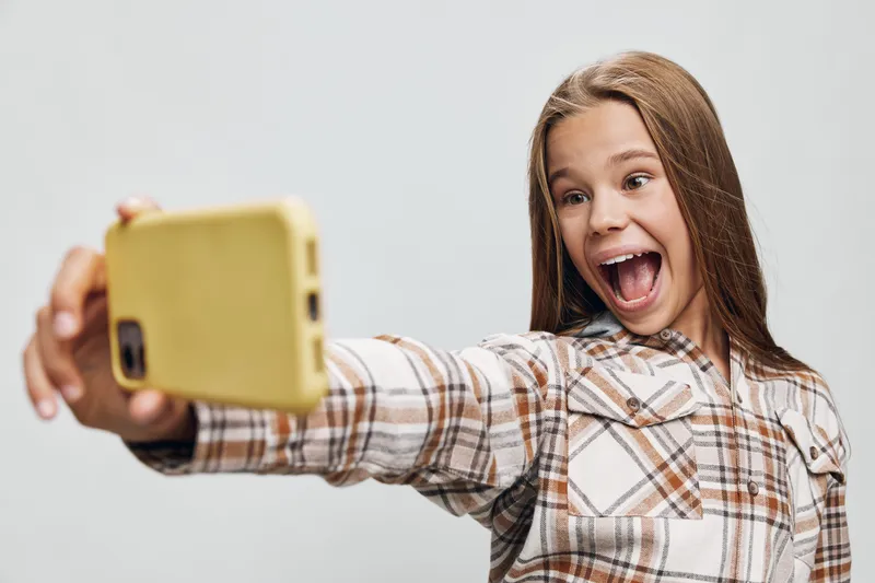 Girl taking selfie with smartphone, smiling and excited. Child wearing casual plaid shirt isolated on gray background. Happy kid capturing photo with mobile phone.