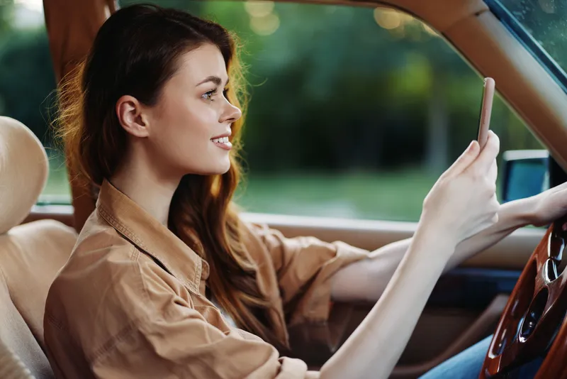 Young woman in a beige shirt driving a car while using her smartphone, joyful expression, nature background, sunny day, the concept of modern lifestyle.