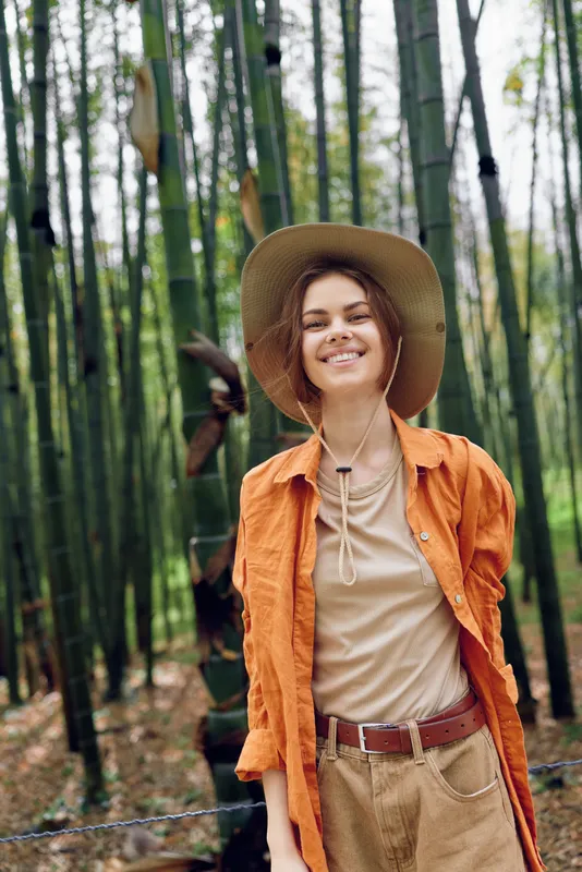 Woman portrait smile hat bamboo forest, young traveler standing in a bamboo grove wearing a wide brim hat and orange shirt, outdoor lifestyle, nature escape, adventure and relaxed mood.