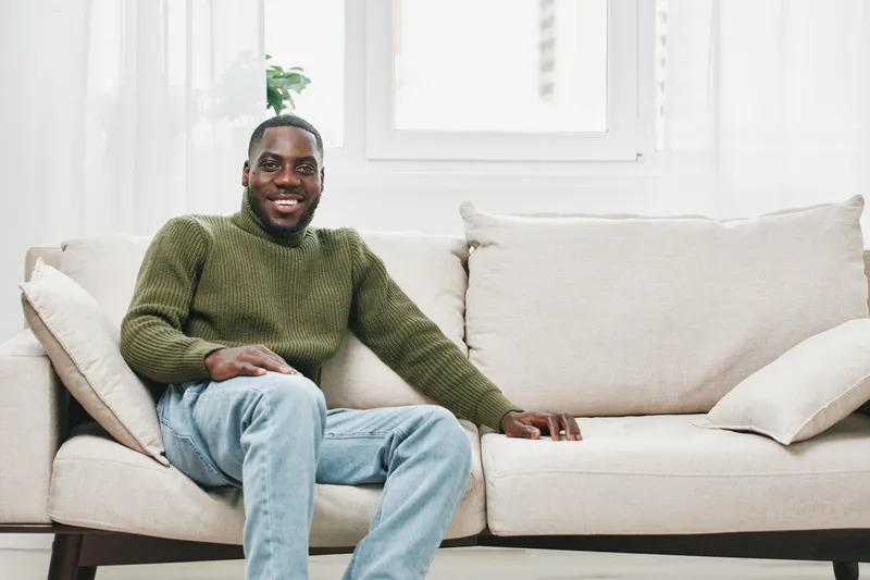 Smiling African American man in a green sweater relaxing on a beige sofa, enjoying a bright and cozy indoor setting, adding warmth and comfort to home decor