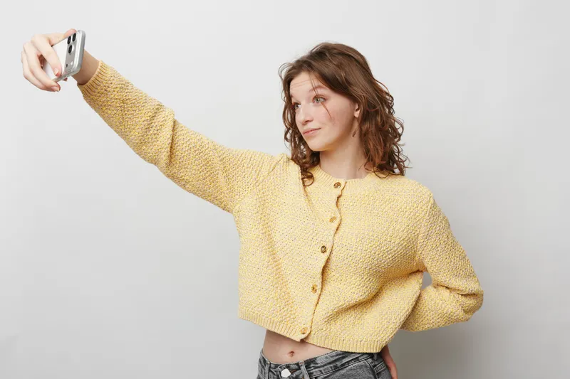 Young woman with curly hair wearing yellow knitted sweater takes selfie with smartphone on grey background. Casual style, social media concept, modern lifestyle and self-expression.