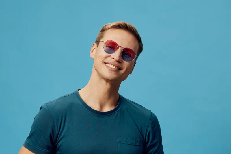 Smiling young man with light skin, wearing blue t-shirt and stylish pink sunglasses, looking happy and confident against a plain bright blue background. Casual fashion, positive emotion, lifestyle con