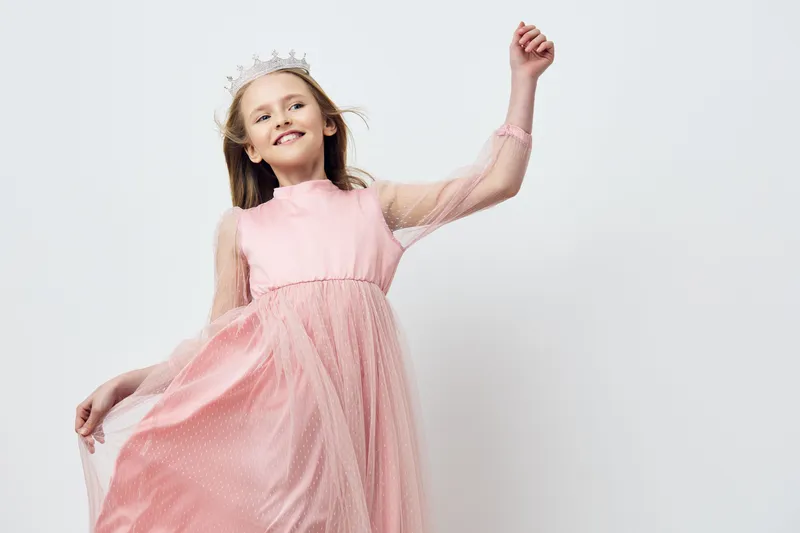 Happy girl in a pink dress twirling joyfully, wearing a crown, on a light background, embodying childhood innocence and joy