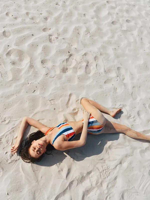 Portrait of a woman sunbathing on white sand, wearing a striped bikini. She lies relaxed with soft shadows, warm light, and a serene seaside mood.