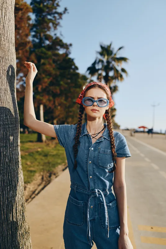 Lifestyle woman in modern boho-western denim street style with sunglasses and braids standing outdoors on a sunny day, film color tone enhancing the vibrant scene.