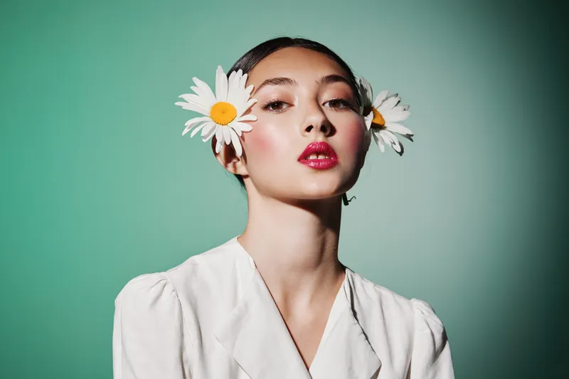 Young woman with nude makeup and shiny lip gloss poses in white garment with daisies in her hair on soft green banner flowing fabric background. Creative beauty and fashion.