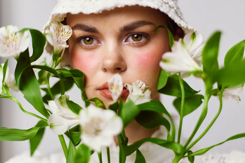 Beautiful young woman with flowers, wearing a hat and soft makeup, showcasing a serene expression in a light, airy setting with a neutral background