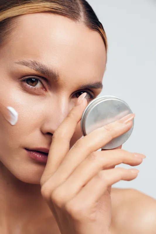 A closeup of a young woman applying face cream using a small round mirror, focusing on skincare and beauty routine in a bright studio setting with natural light.