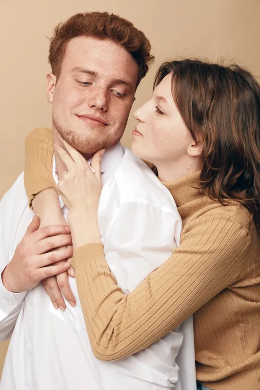 Young couple embraces tenderly, woman in beige sweater hugs man in white shirt. Intimate moment of affection and love captured in warm neutral background studio setting.