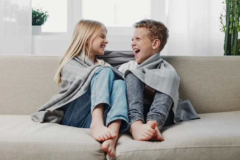 Happy children wrapped in grey blanket laughing together on beige sofa in bright room. Cozy family moment shows warmth, joy, sibling bond, and playful indoor lifestyle concept.