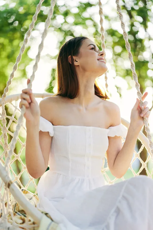 Young woman enjoying serene moments on a swing in a sunlit garden, wearing a white dress, exuding happiness and tranquility in a nature setting