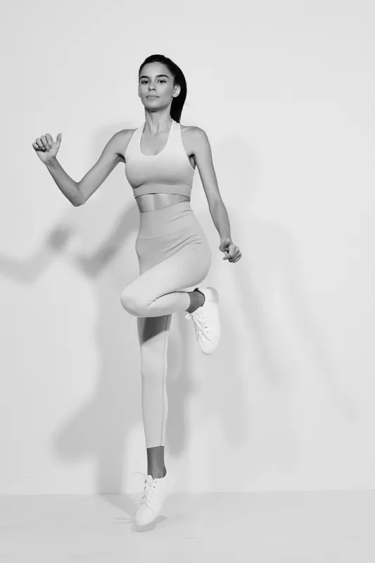 Woman sport fitness dressed in light activewear performing high knee exercise indoors, showing strength, balance, and focus in black and white studio setting.