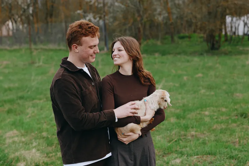 Cheerful couple outdoors, smiling at each other, softly holding a small dog, sunlit scene, autumnal colors, casual outfits, relaxed posture, gentle breeze, lush greenery background, natural warmth,