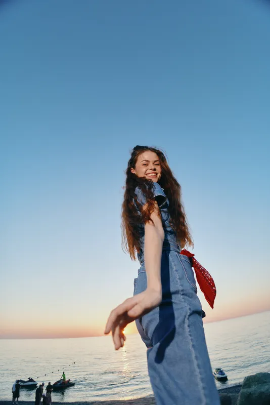 Lifestyle woman in modern boho-western denim street style with a film color effect enjoys coastal sunset, smiling with carefree energy and natural beauty by the sea.