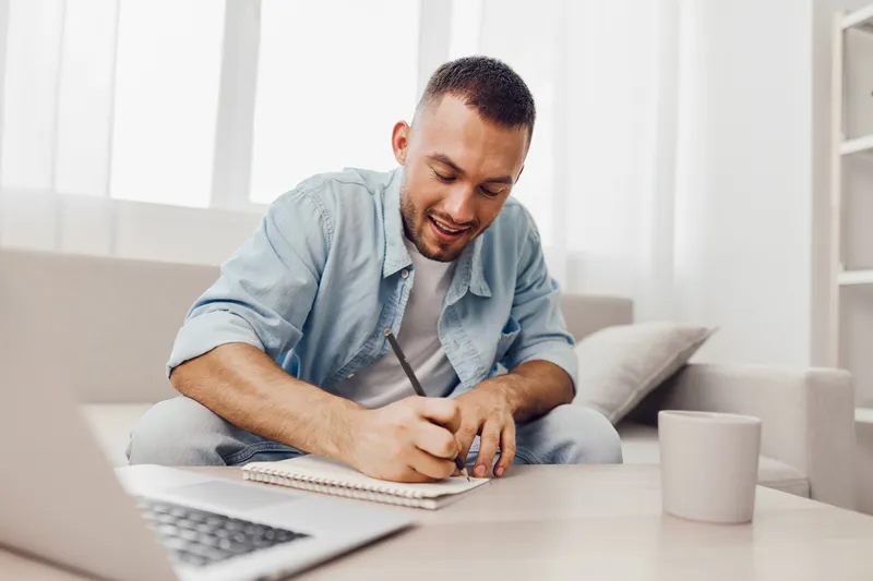 Relaxed man writing in a notebook at home, showcasing creativity and focus on a soft, cozy couch. Ideal for themes of remote work and self-improvement.