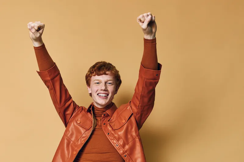 Young man with red hair raising hands in celebration and smiling wearing orange shirt and jacket against beige background for success and happiness.