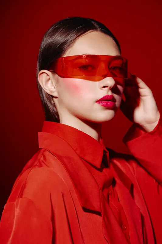 Young woman in a stylish red outfit wearing futuristic red transparent sunglasses poses against a red background. Fashion and beauty editorial concept with creative vibes.