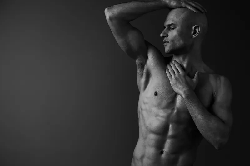Black and white image of a shirtless man posing confidently, showcasing his muscles and striking a powerful yet contemplative expression.