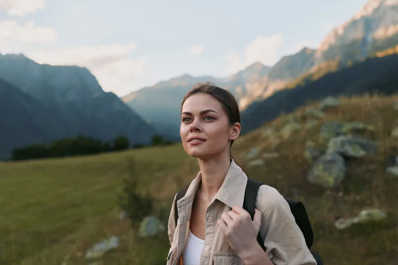 Woman traveler hikes through a sunny meadow with a backpack, enjoying nature and dramatic mountains, a candid outdoor lifestyle moment amid expansive scenery and peaceful solitude outside.