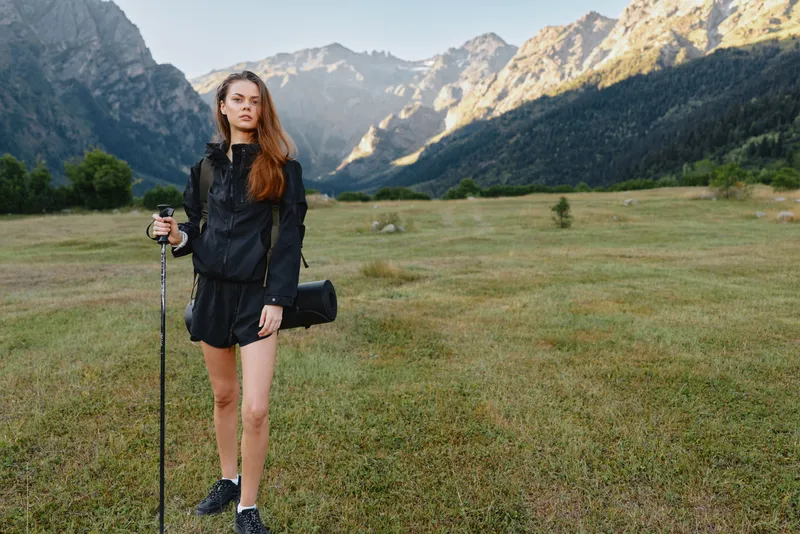 A young woman stands in a mountain meadow with a trekking pole, wearing dark outdoor gear and a crossbody bag. She appears confident and ready for an adventurous outdoor day.