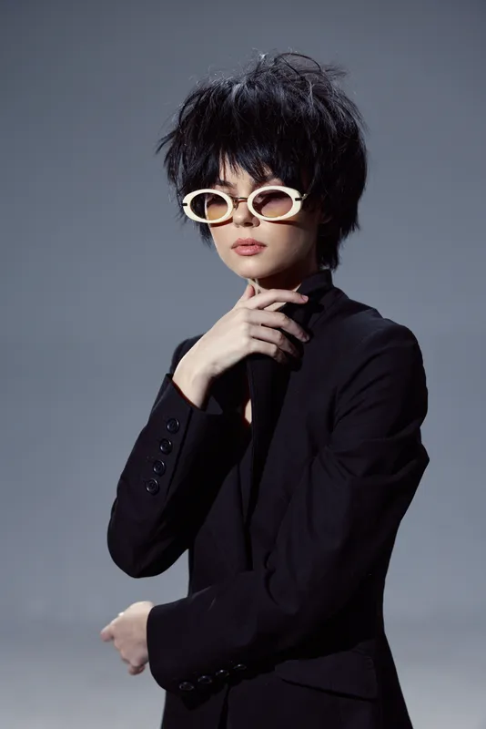 Stylish woman with short black hair and white sunglasses wearing a black blazer posing confidently against a grey background.