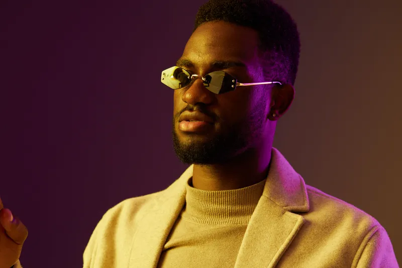 Stylish young man with sunglasses in a cream colored coat against a vibrant purple background, conveying confidence and modern fashion trends