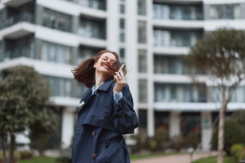 Young woman with long hair wearing a dark trench coat smiles joyfully while holding sunglasses outdoors in an urban environment during daytime.