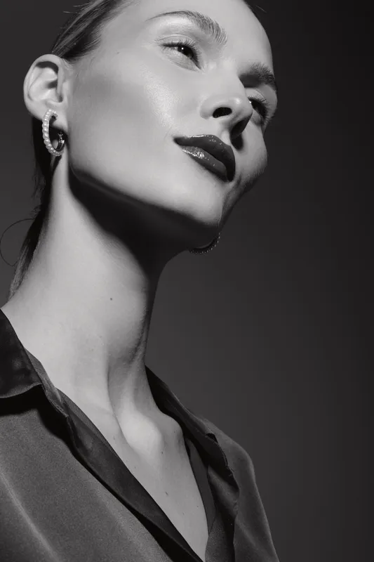 Close-up portrait of a young white woman with fair skin and glossy lips wearing earrings and a silky top. The black and white photo highlights elegance and fashion style.