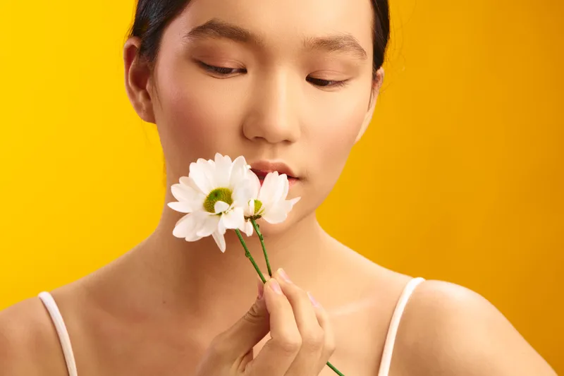 Asian woman gently holds a white flower near her lips against a bright yellow backdrop, conveying calm elegance and mindful beauty in a soft studio mood.