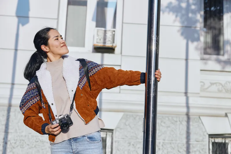 Woman with camera leaning against a pole outdoors, wearing a colorful jacket and jeans, smiling as she photographs a street scene. Casual, confident, capturing candid moments in daylight.