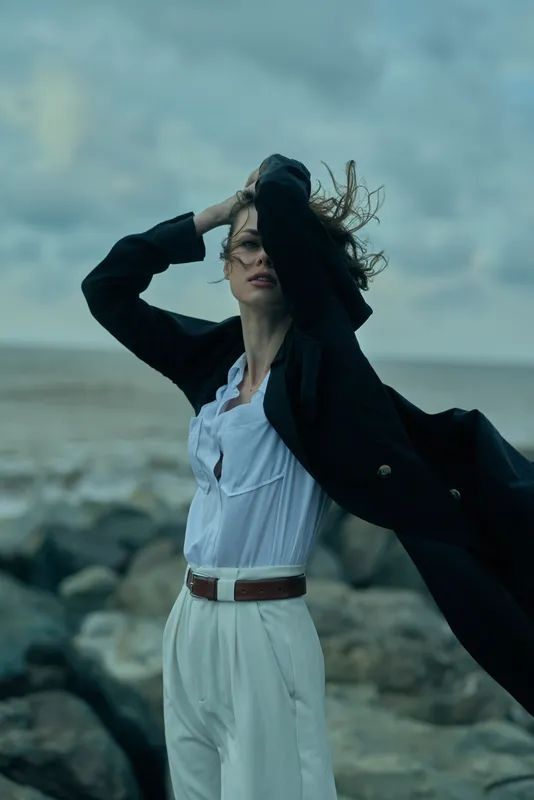 Wind swept coat and light dress by the sea, dynamic movement under a moody sky, fashion photography with outdoor breeze and dramatic ocean backdrop