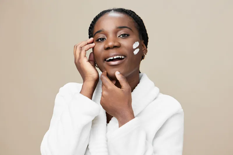 Young African woman applying facial cream, smiling and looking at the camera, wearing a white robe against a soft beige background