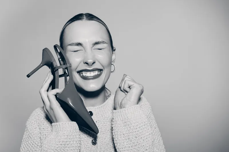 Happy caucasian woman with fair skin holding high heel shoe near face smiling with eyes closed in cozy sweater. Concept of joy, fashion, and female lifestyle.