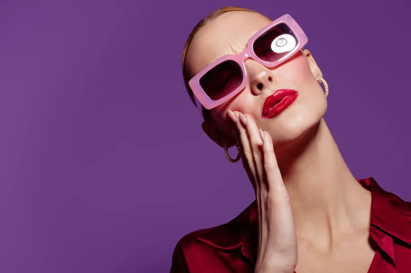 Stylish woman with pink sunglasses and red lipstick poses with hand on face against purple background. Fashion portrait with bold makeup and trendy accessories concept.