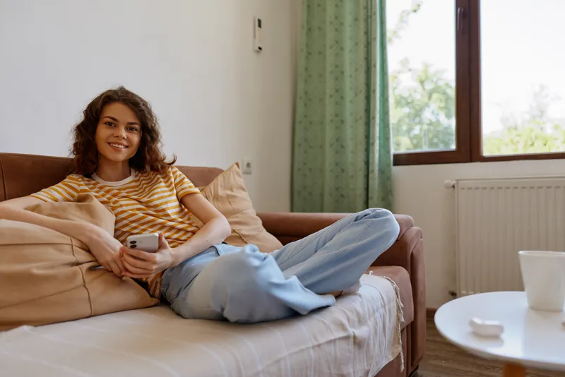 A young woman with curly hair sits on a sofa at home, smiling while scrolling her phone, creating a relaxed, everyday lifestyle moment on a comfortable couch.