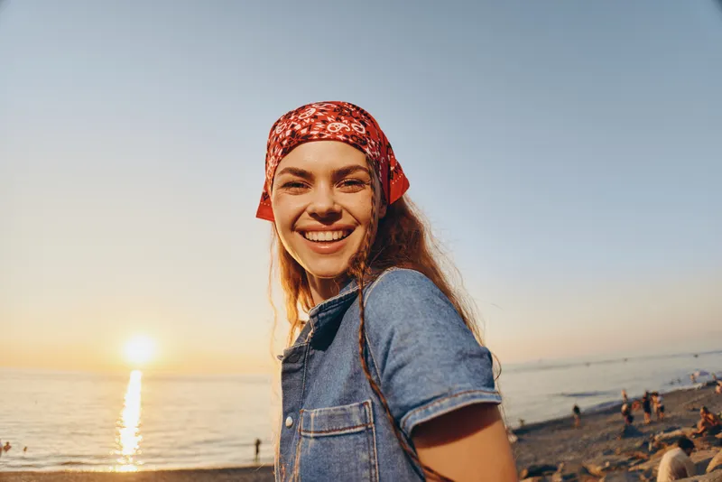 smiling woman in modern boho-western denim street style with film color effect at sunset on beach, natural light, casual joy, carefree lifestyle