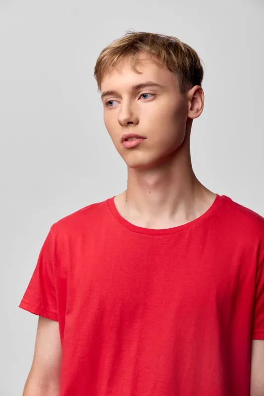 Young man in a red t shirt stands against a neutral backdrop, relaxed pose and soft lighting, showcasing casual style and contemporary fashion for editorial stock and lifestyle imagery